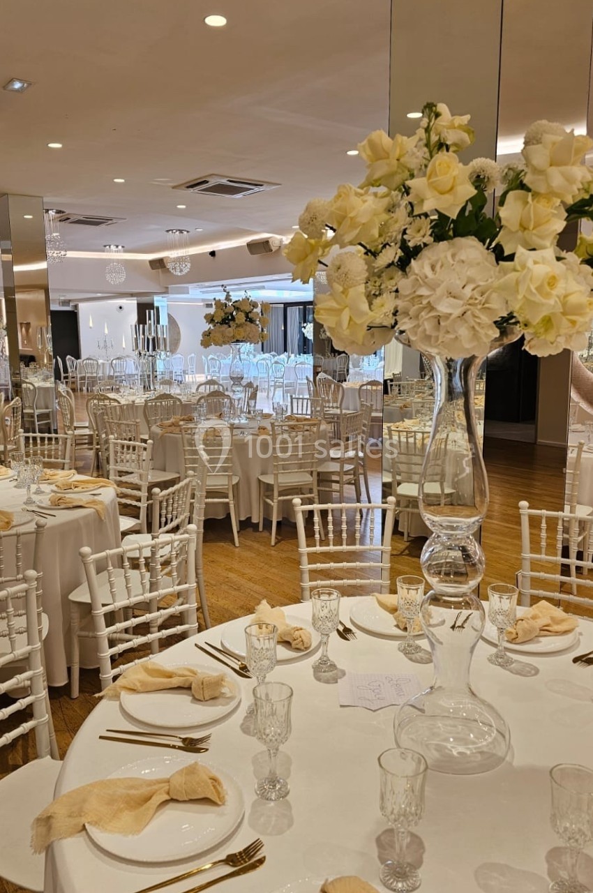 Salle de réception élégante avec tables rondes dressées, nappes blanches, chaises blanches et grands bouquets de fleurs.
