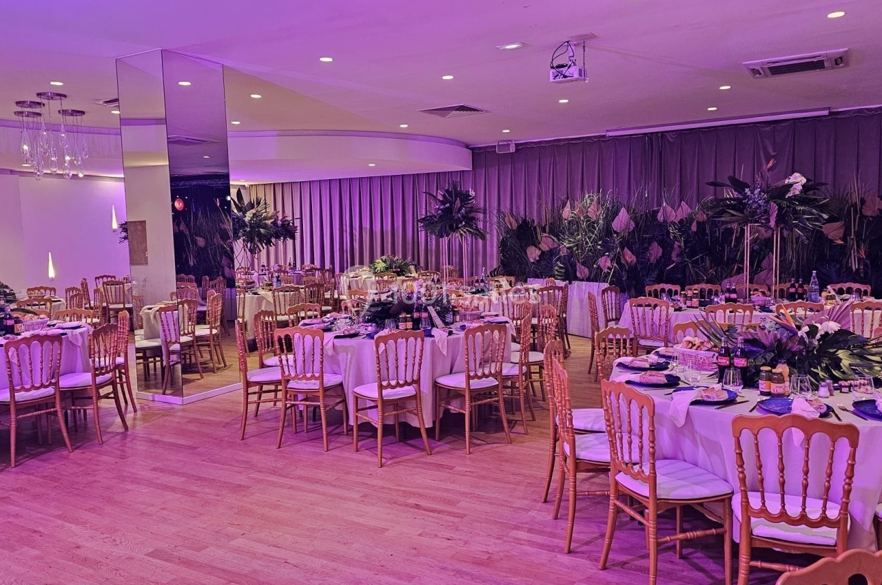 Salle de réception décorée avec des tables rondes dressées, des chaises en bois et un éclairage violet tamisé.