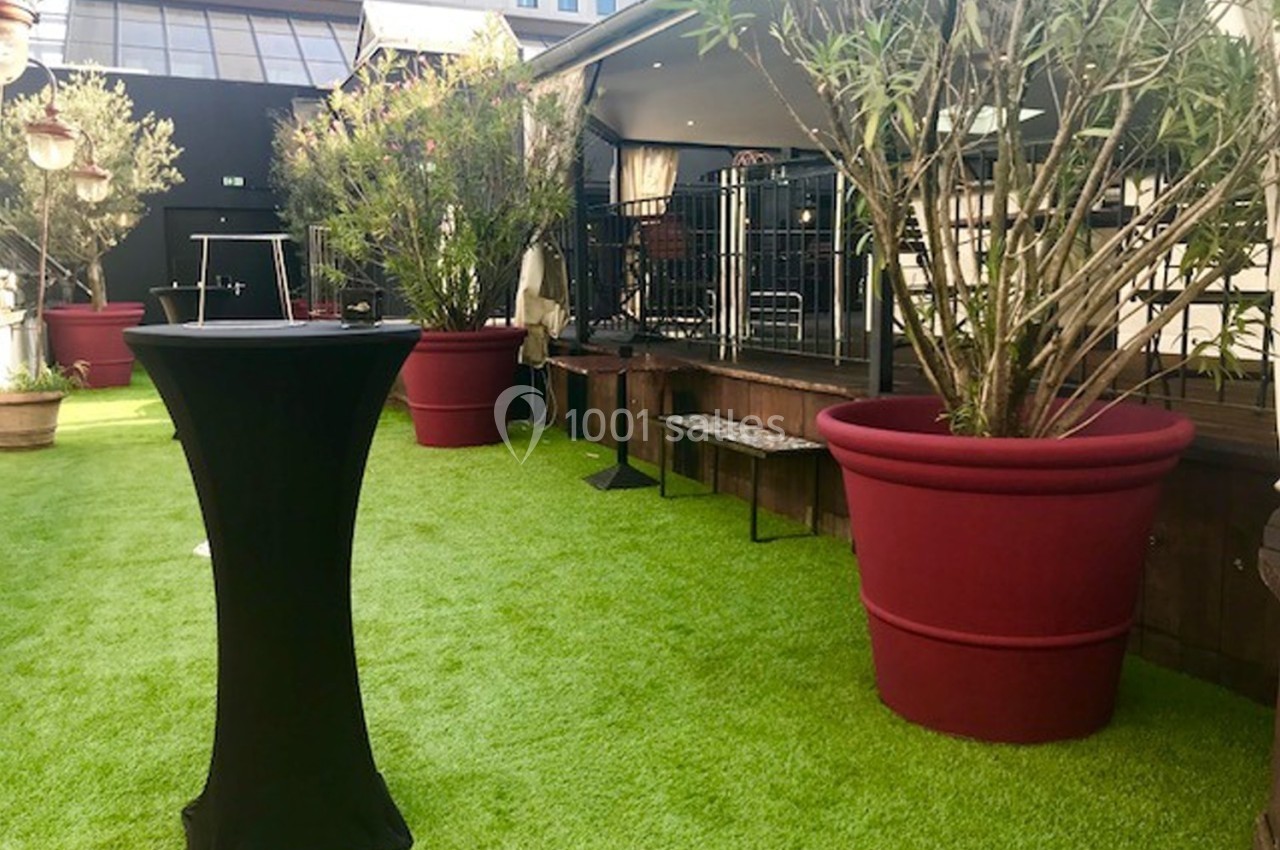Terrasse avec gazon artificiel, grandes plantes en pots rouges et mobilier de réception près d'une estrade.
