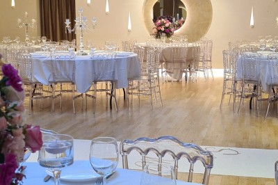 Salle de réception élégante avec tables rondes dressées, chaises transparentes et décorations florales.