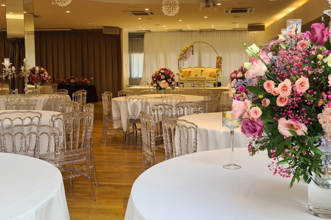 Salle de réception décorée avec des tables rondes, nappes blanches et grands bouquets de fleurs colorées.