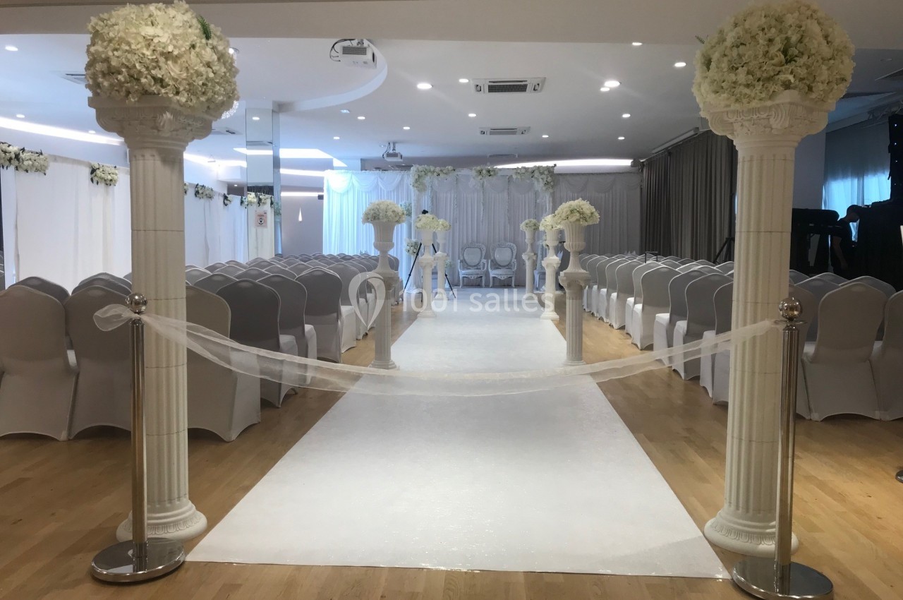 Salle décorée pour une cérémonie avec allée centrale bordée de colonnes blanches ornées de fleurs et chaises alignées.