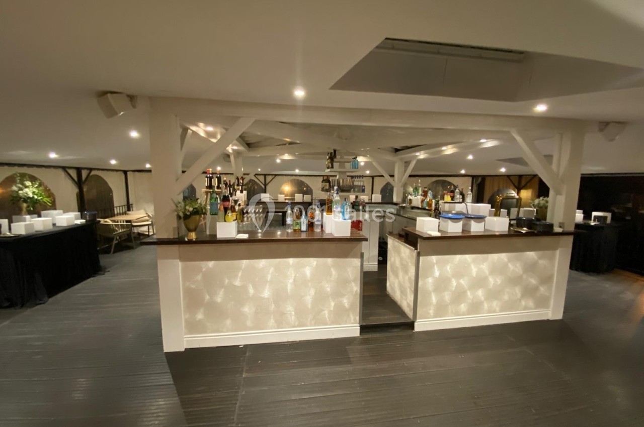Bar central moderne avec comptoir en bois sombre, étagères de bouteilles et éclairage tamisé dans une grande salle.