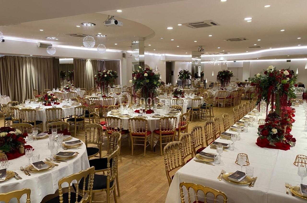 Salle de réception élégamment décorée avec des tables dressées, des chaises dorées et des arrangements floraux rouges.