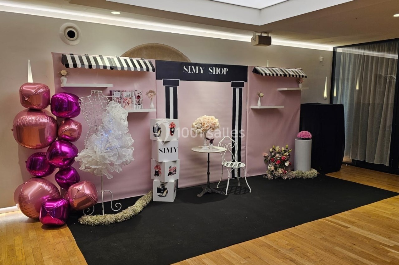 Décor rose avec ballons, présentoirs, robe blanche, table et chaise, évoquant une boutique ou un espace d'exposition.