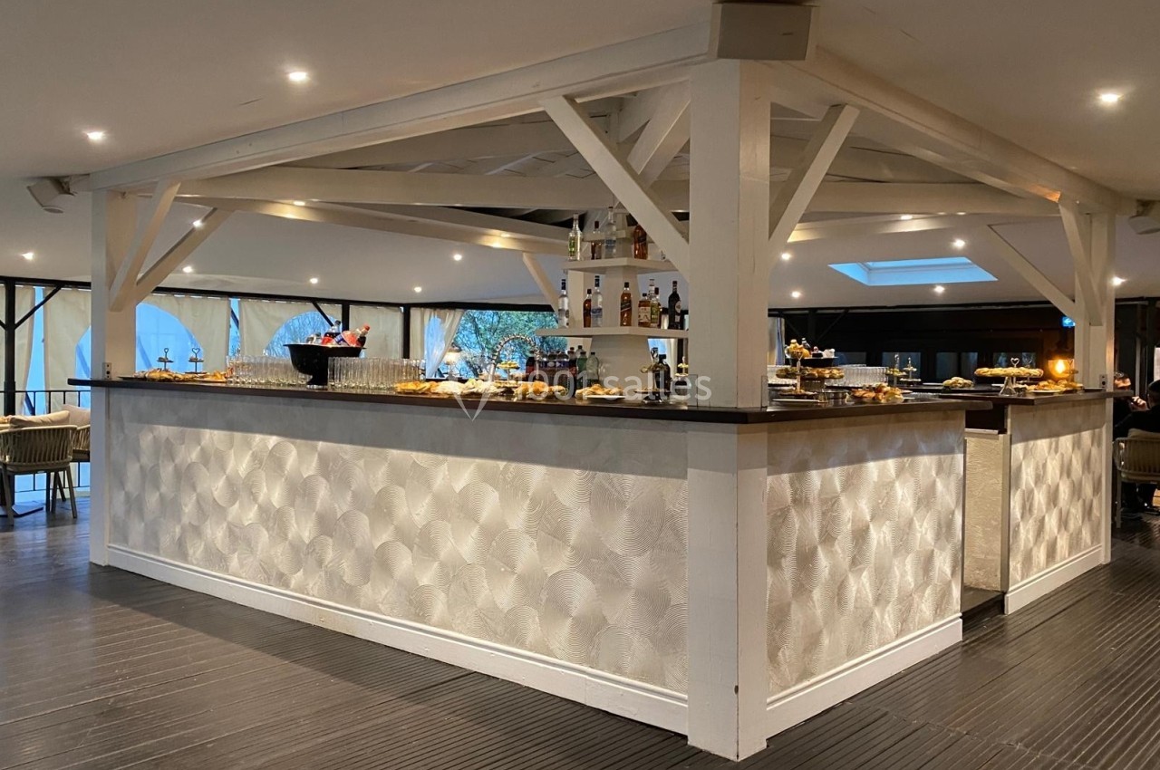 Bar moderne en bois clair avec comptoir texturé, entouré de tables et décoré de bouteilles et plats.