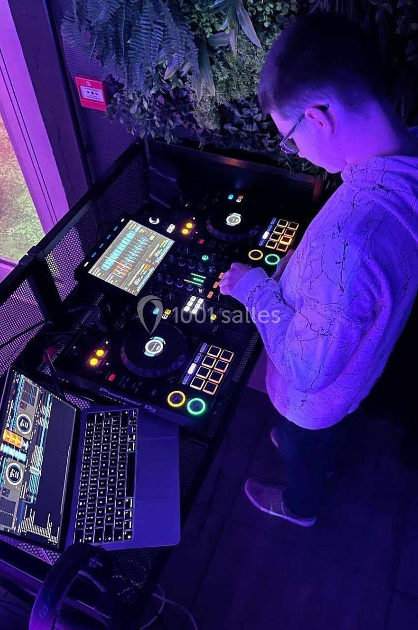 Un DJ mixe de la musique sur une console équipée d'écrans et de lumières colorées, avec un ordinateur portable à côté.