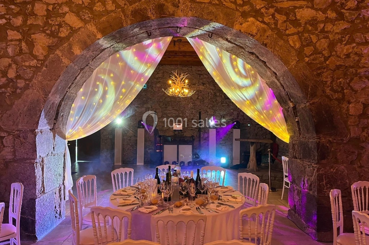 Salle en pierre décorée pour un événement, avec une table ronde dressée et des éclairages colorés sous une arche.