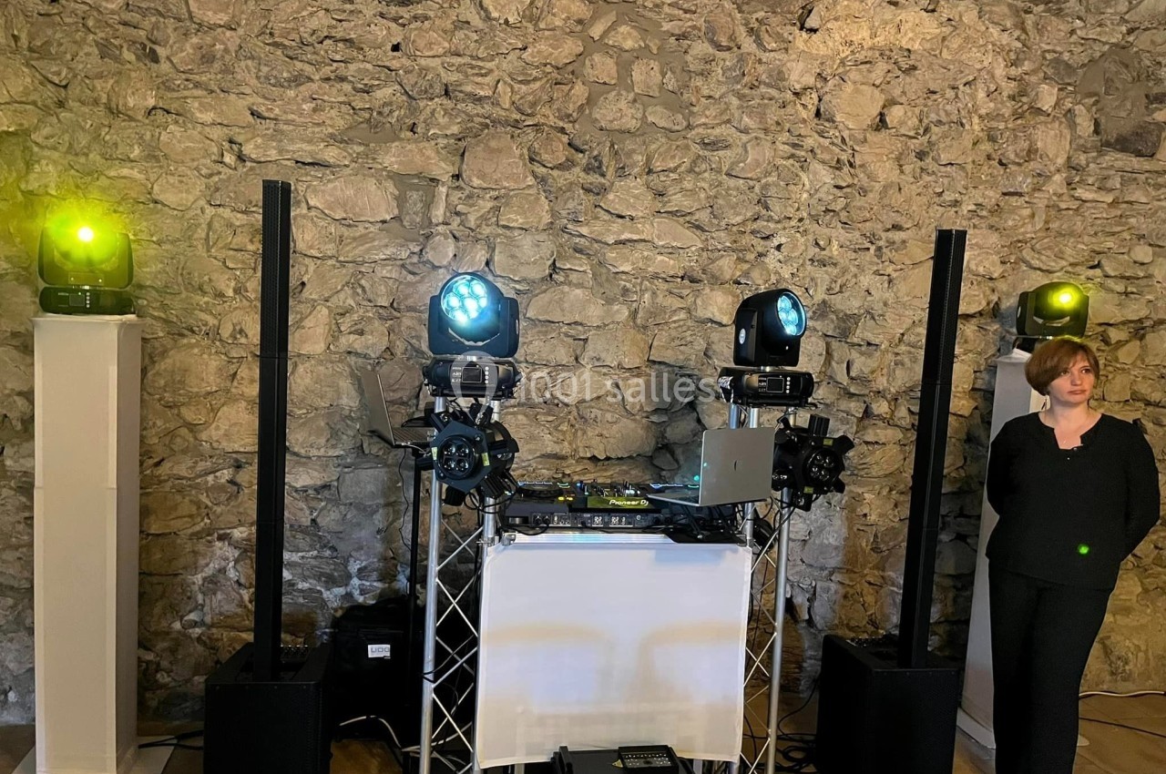 Installation de matériel de sonorisation et d'éclairage devant un mur en pierre, avec une personne debout à droite.