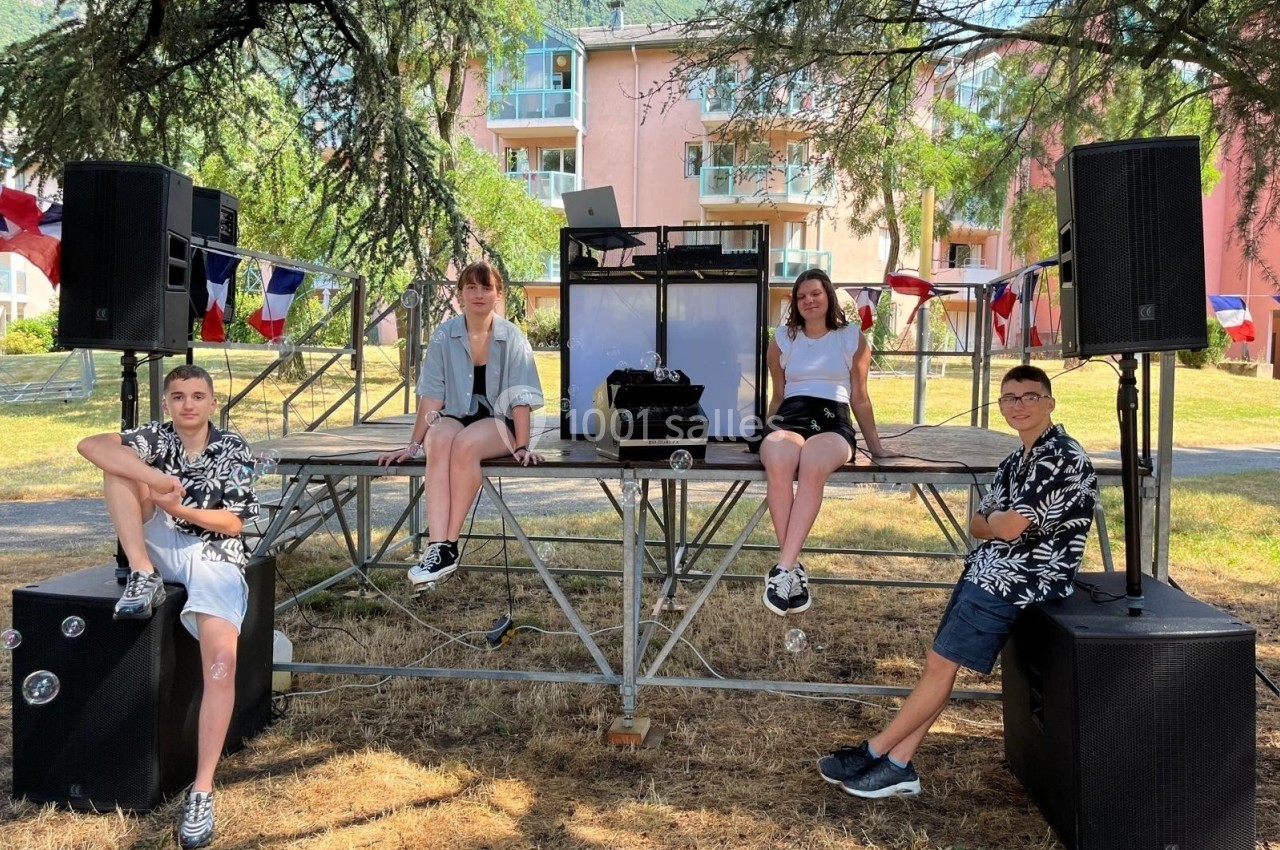 Quatre jeunes posent sur une scène extérieure avec des équipements audio, entourés de décorations et de drapeaux français.