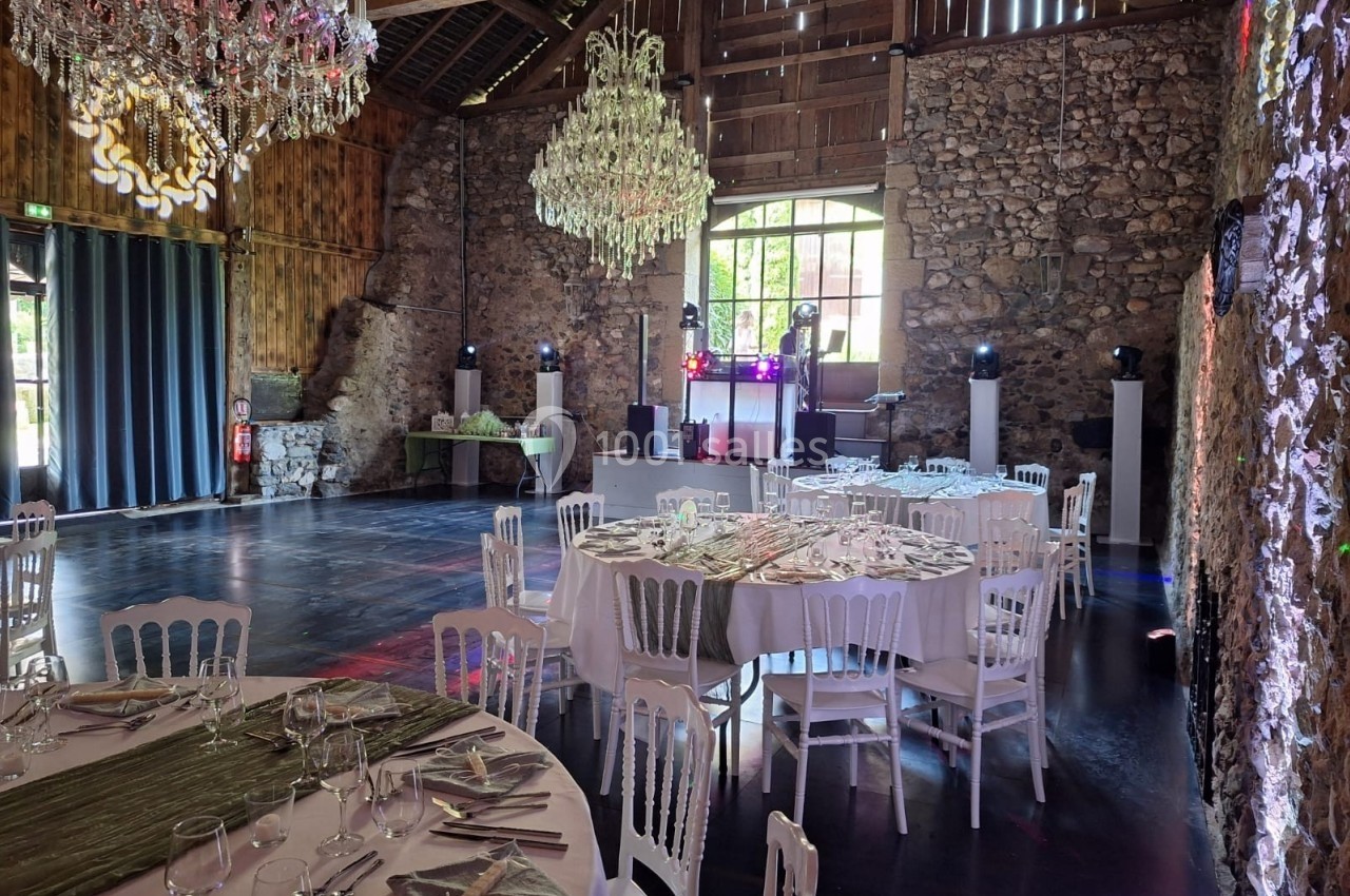 Salle de réception rustique avec murs en pierre, tables dressées, chaises blanches et lustres suspendus.