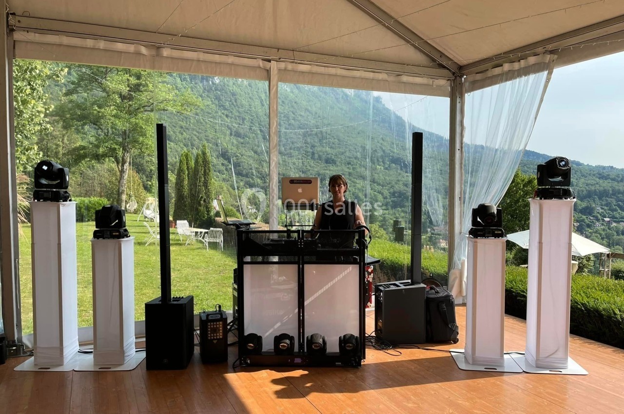 Un DJ mixe sous une tente ouverte avec vue sur un paysage verdoyant et des équipements de sonorisation.