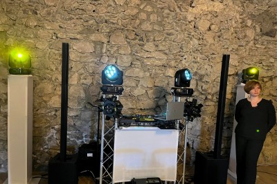 Installation de matériel de sonorisation et d'éclairage devant un mur en pierre, avec une personne debout à droite.