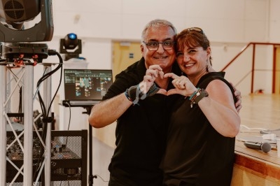 Un homme et une femme souriants forment un cœur avec leurs mains dans une salle équipée de matériel audiovisuel.