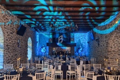 Salle en pierre avec des tables dressées, chaises blanches, éclairage bleu et motifs lumineux au plafond en bois.