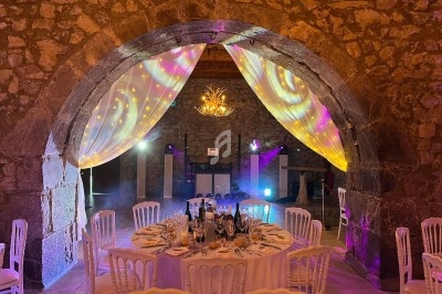 Salle en pierre avec une table ronde dressée pour un repas, éclairée par des lumières colorées et des voilages suspendus.