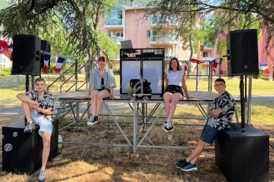 Quatre jeunes posent sur une scène extérieure équipée de matériel audio, entourée de drapeaux français.