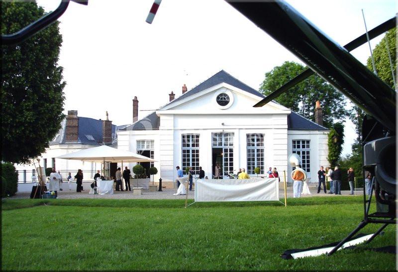 Façade d'un bâtiment élégant avec des invités rassemblés dans un jardin, vue partielle d'un hélicoptère au premier plan.