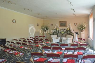 Salle décorée pour un événement avec un piano au premier plan, des chaises rouges alignées et des arrangements floraux.
