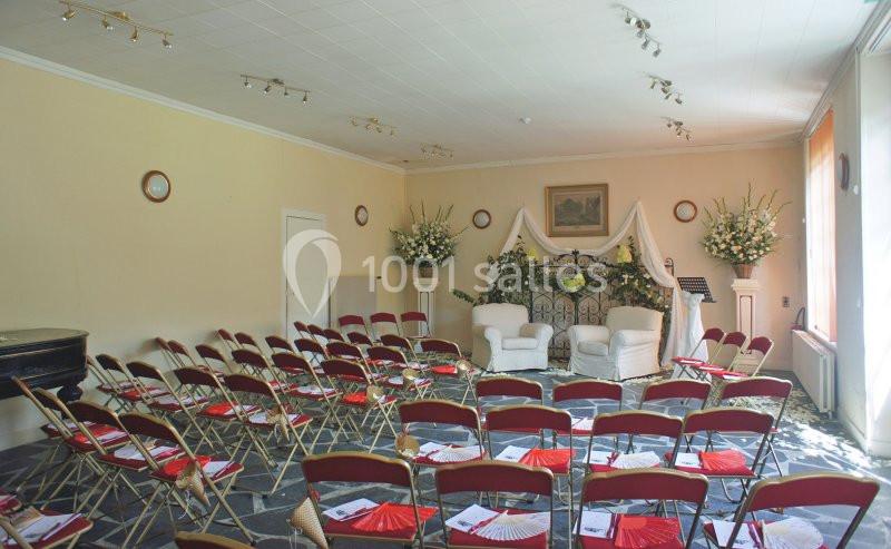 Salle lumineuse aménagée pour un événement avec des chaises pliantes rouges, une arche décorée et des fleurs blanches.