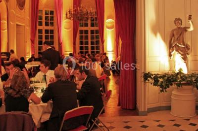 Salle décorée pour un événement avec un piano au premier plan, des chaises rouges alignées et des arrangements floraux.