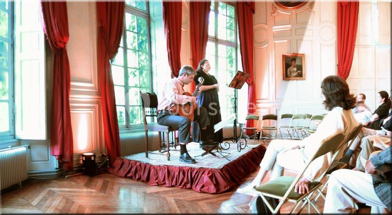 Deux musiciens jouent sur une petite scène dans une salle élégante avec des rideaux rouges et un public assis.