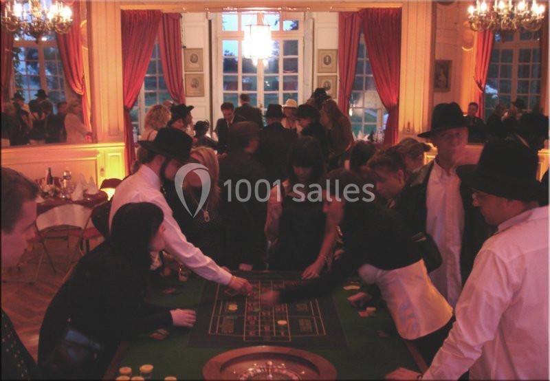 Des personnes jouent autour d'une table de casino dans une salle éclairée avec des rideaux rouges et des lustres.