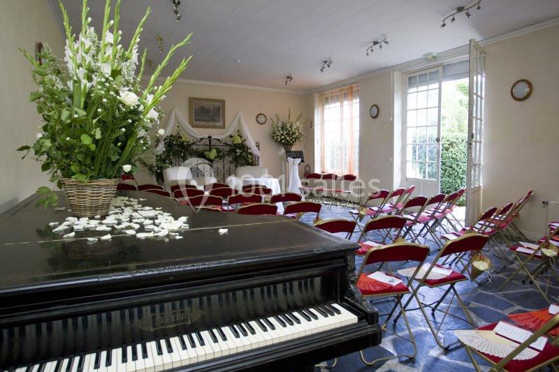 Salle décorée pour un événement avec un piano au premier plan, des chaises rouges alignées et des arrangements floraux.
