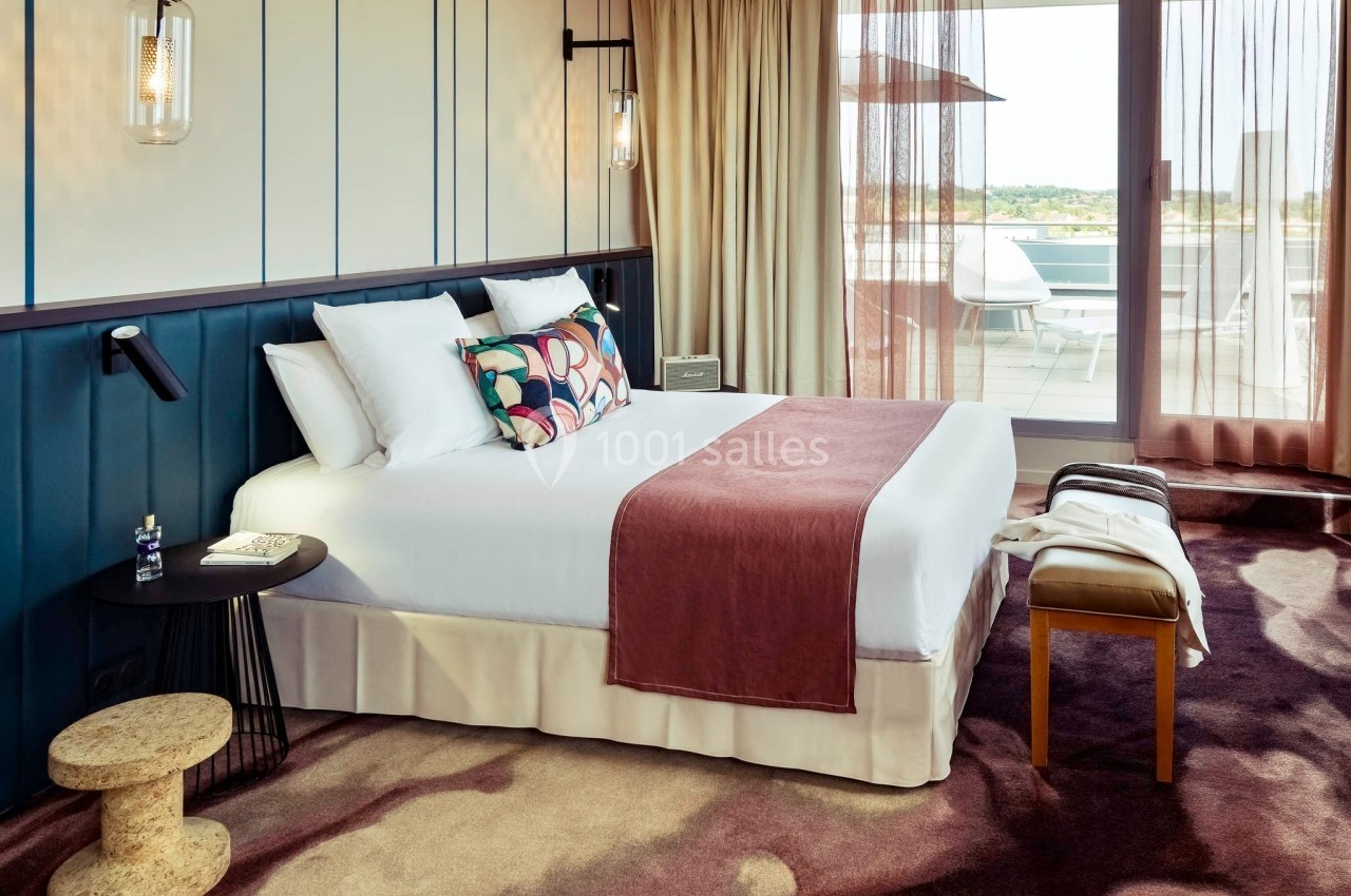 Chambre d'hôtel lumineuse avec lit double, plaid rose, table d'appoint et balcon donnant sur une terrasse extérieure.