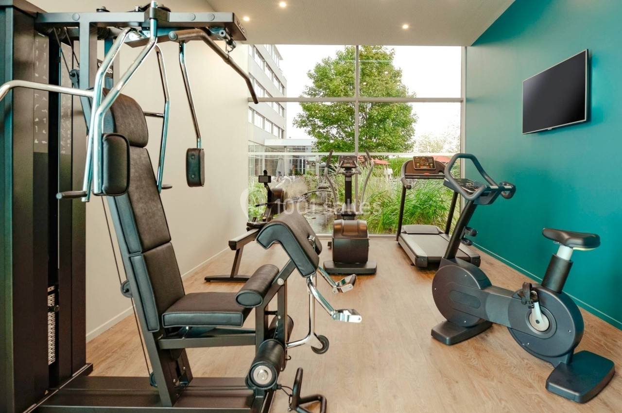 Salle de sport lumineuse avec équipements de musculation, vélo d'appartement, tapis de course et vue sur un jardin.