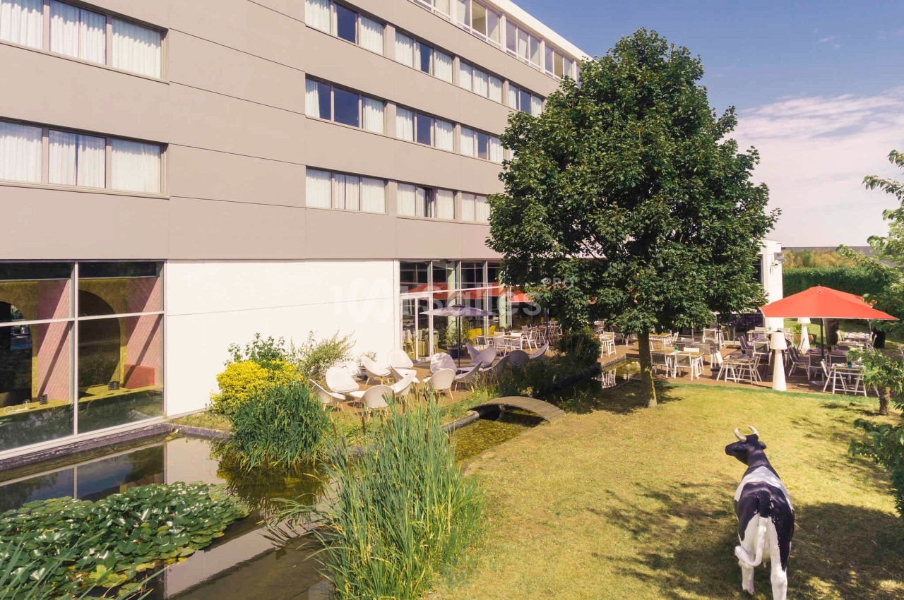 Façade d'un bâtiment moderne avec terrasse, parasols rouges, jardin paysager, étang et statue de vache.