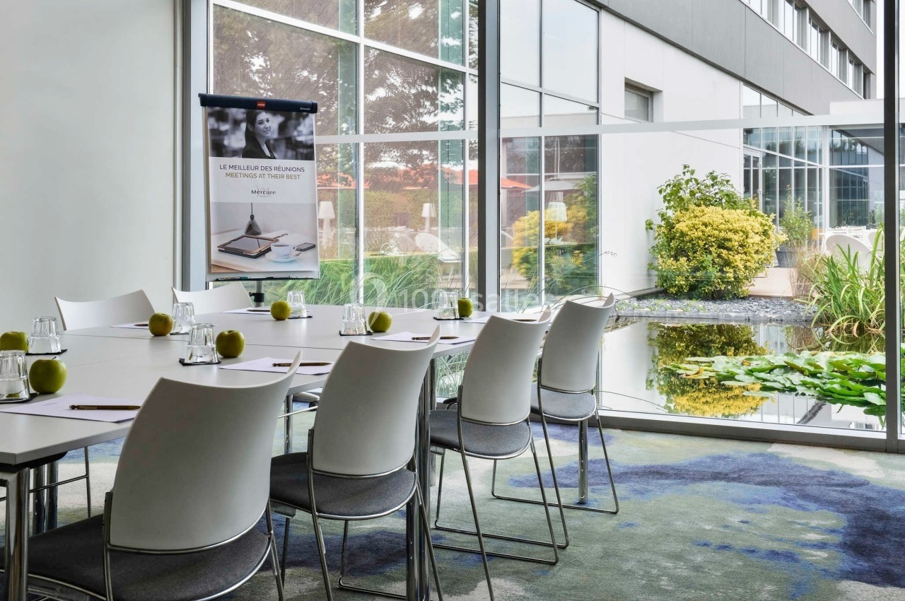 Salle de réunion lumineuse avec table rectangulaire, chaises blanches, pommes vertes et vue sur un jardin extérieur.