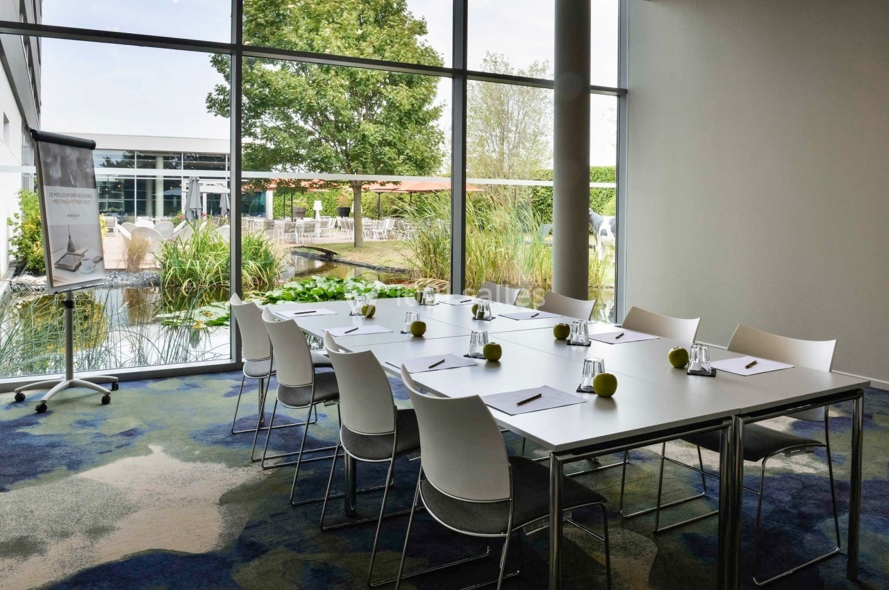 Salle de réunion lumineuse avec une grande table, des chaises blanches, des blocs-notes et vue sur un jardin extérieur.