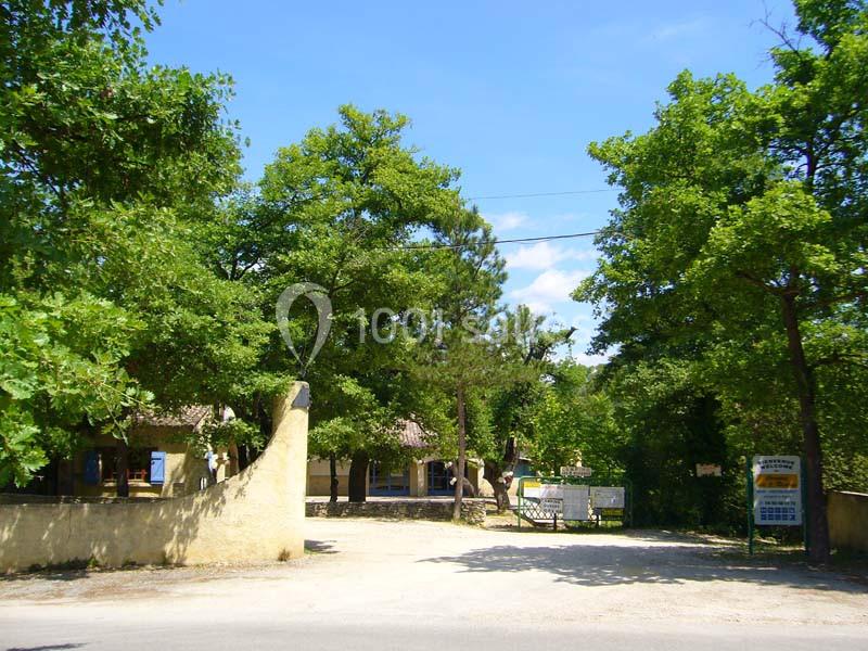 Entrée d'un domaine entourée d'arbres, avec un portail ouvert et des bâtiments visibles en arrière-plan.