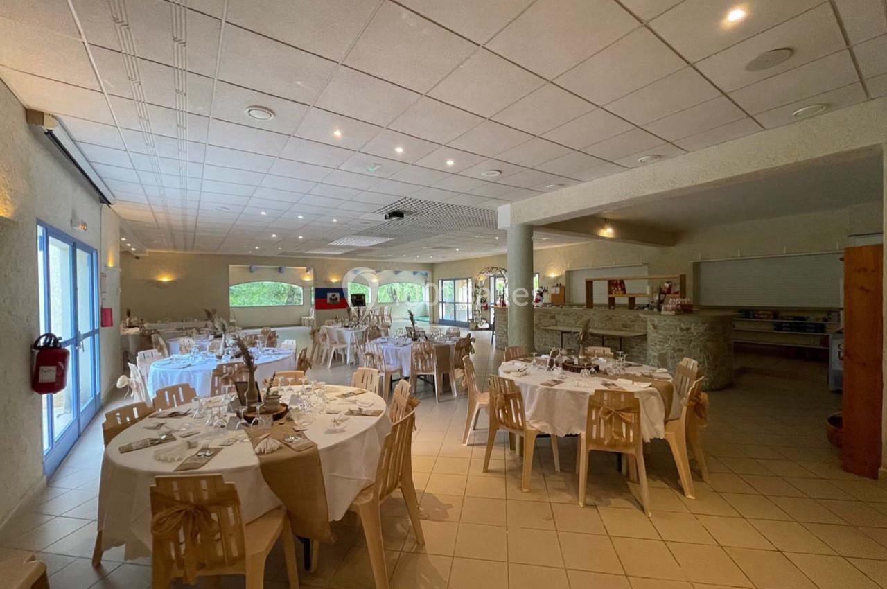 Salle de réception lumineuse avec tables rondes dressées pour un repas, décorée de nappes blanches et chaises en bois.