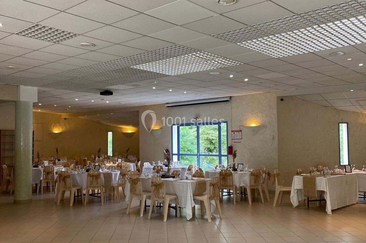 Salle de réception lumineuse avec tables dressées, nappes blanches et chaises décorées, vue sur un espace extérieur…