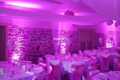 Salle de réception décorée avec des nappes blanches, des chaises ornées de rubans roses et un éclairage violet.