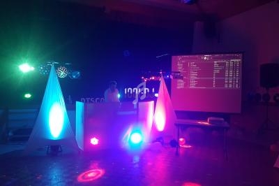 Salle sombre avec un DJ derrière une table éclairée par des lumières colorées et un écran projetant une playlist.