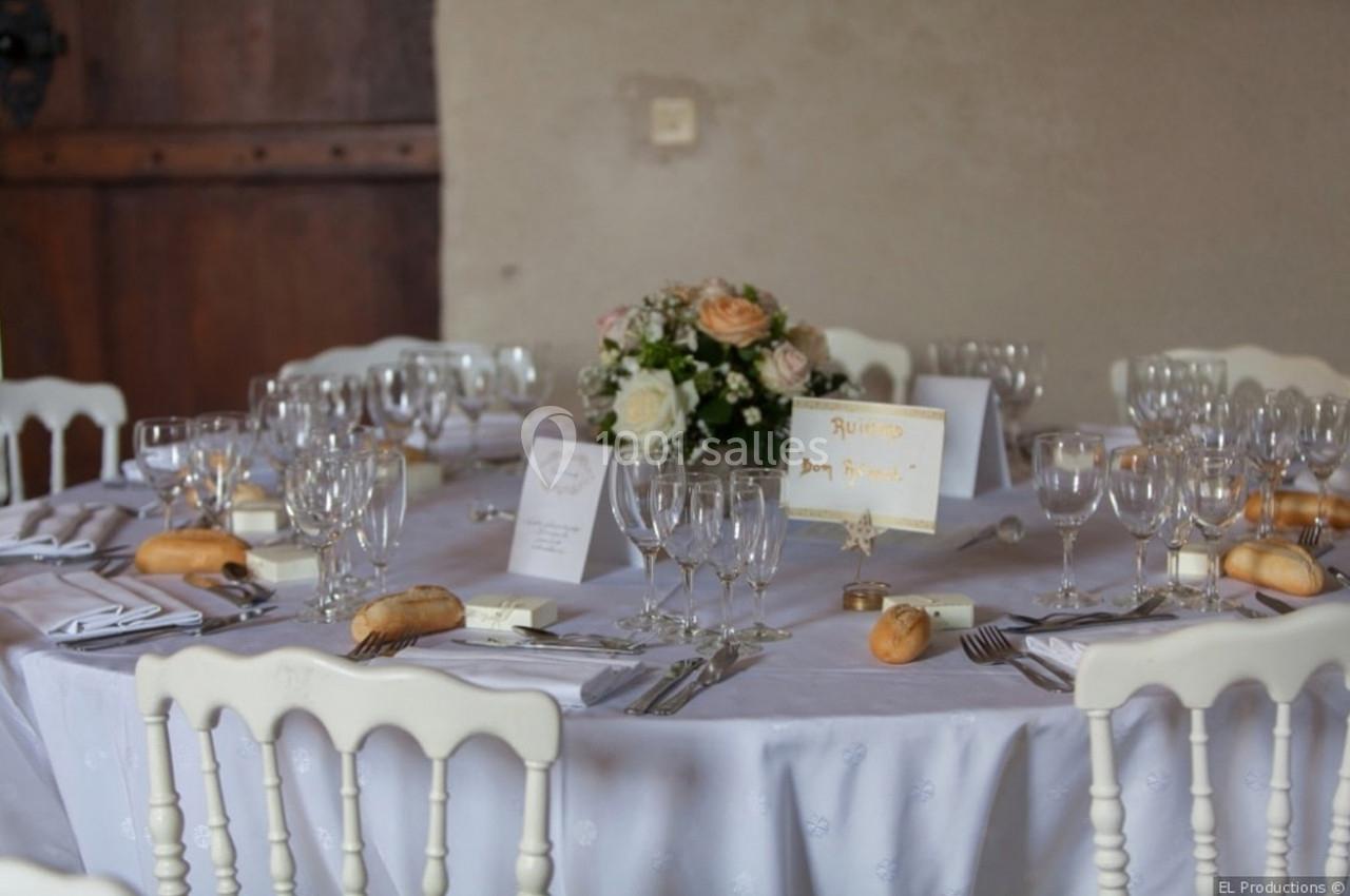 Table ronde dressée pour un repas, avec nappes blanches, verres, menus, fleurs au centre et petits pains.