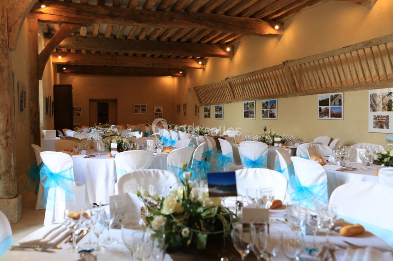 Salle de réception décorée avec des tables rondes, nappes blanches, chaises ornées de rubans bleus et compositions florales.