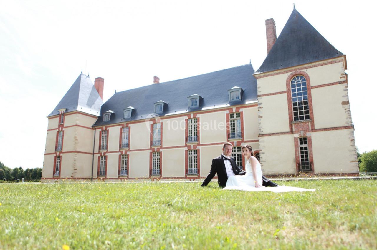 Un couple en tenue de mariage assis sur une pelouse devant un château à l'architecture classique.