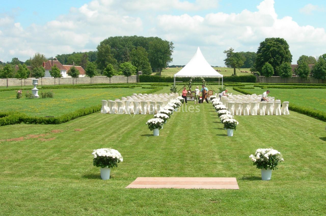 Chaises blanches alignées sur une pelouse avec une tente blanche au fond, entourées de fleurs et d'arbres.