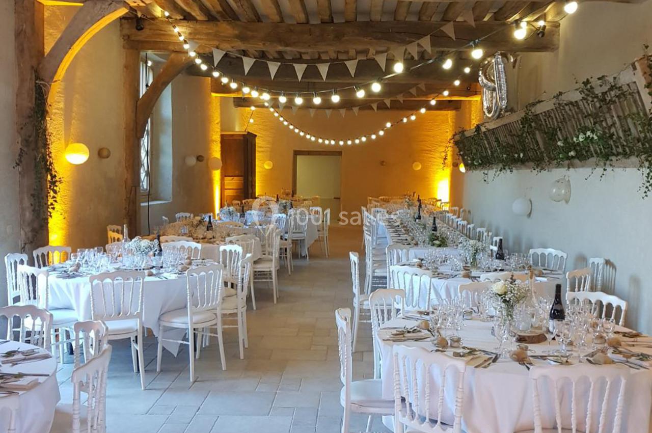 Salle de réception décorée avec des tables rondes, des chaises blanches, des guirlandes lumineuses et des éléments végétaux.