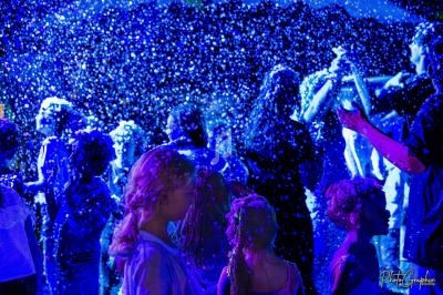 Des enfants et adultes jouent sous une pluie de mousse illuminée par des lumières bleues dans une ambiance festive.