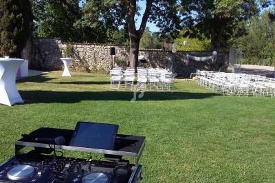 Tables hautes, chaises blanches et matériel de DJ installés dans un jardin pour un événement en plein air.