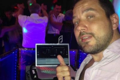 Un homme devant un ordinateur portable, entouré de personnes dansant dans une ambiance festive et éclairée.