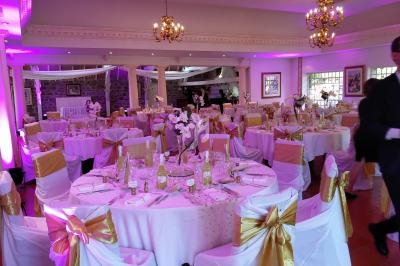 Salle de réception décorée avec des tables rondes, nappes blanches, chaises ornées de rubans dorés et éclairage rose.
