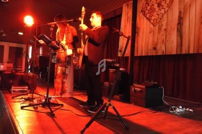 Un duo de musiciens joue sur une scène éclairée en rouge, avec un décor en bois et un cœur mural en arrière-plan.