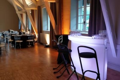 Salle de réception avec tables dressées, bar lumineux blanc et tabourets noirs sur un sol en parquet.