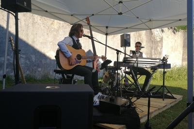 Deux musiciens jouent sous une tente blanche, l'un à la guitare et l'autre au clavier, avec des équipements audio autour.